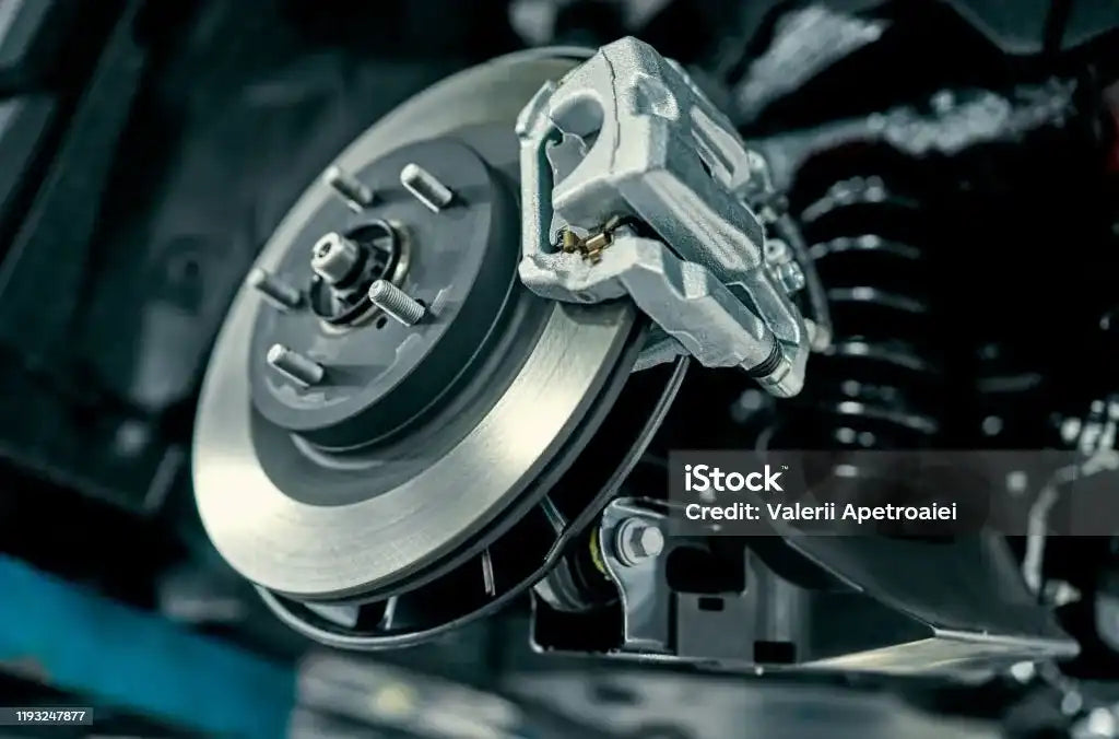 Close-up of a car brake assembly featuring a silver metal brake rotor and caliper, with a dark engine background. 