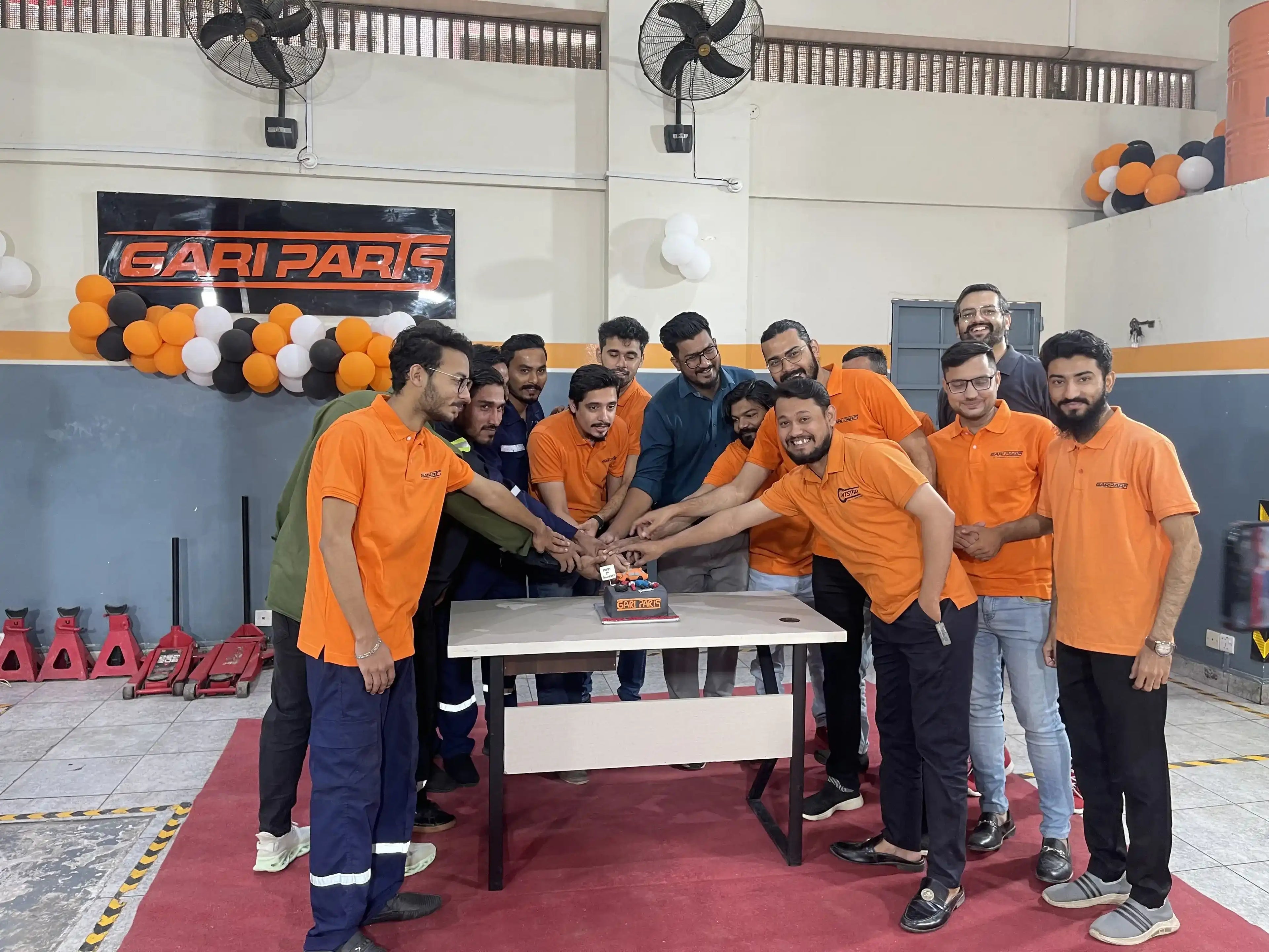 A group of men in matching orange polo shirts gather around a small cake on a table inside a garage or workshop space. A large 'GARI PARIS' sign is visible on the wall behind them, along with orange, black, and white balloon decorations.