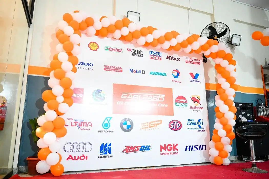 A large white sponsor backdrop banner decorated with an orange and white balloon arch, featuring numerous automotive and brand logos including Shell, Castrol, Suzuki, Honda, Audi, BMW, and NGK, set in an indoor event space.